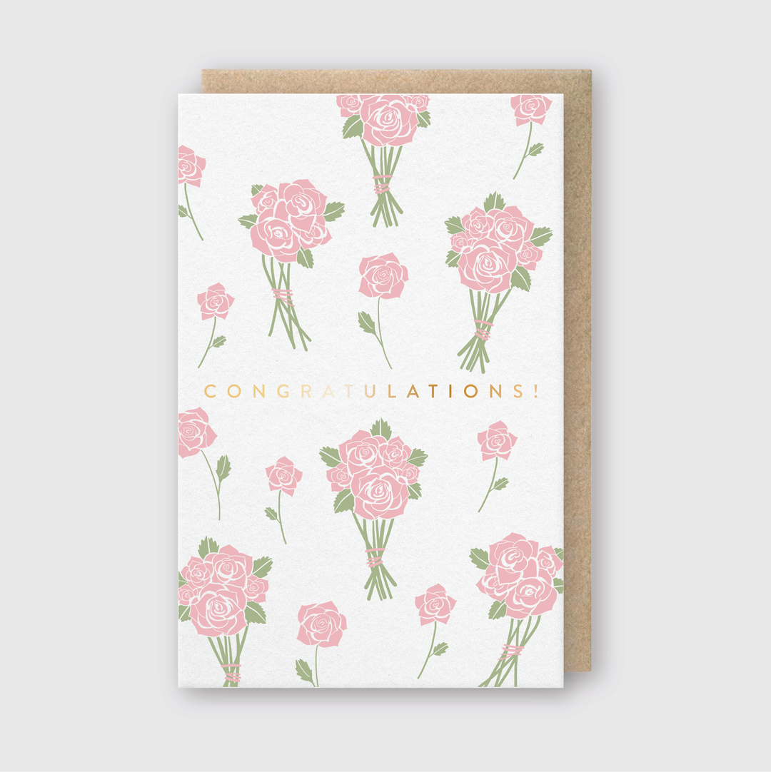 The Congrats Flower Bunches Greeting Card showcases pink rose bouquets elegantly arranged across a white backdrop. At the center, "Congratulations!" is prominently inscribed in gold capital letters. The card comes with a discreetly positioned kraft paper envelope.