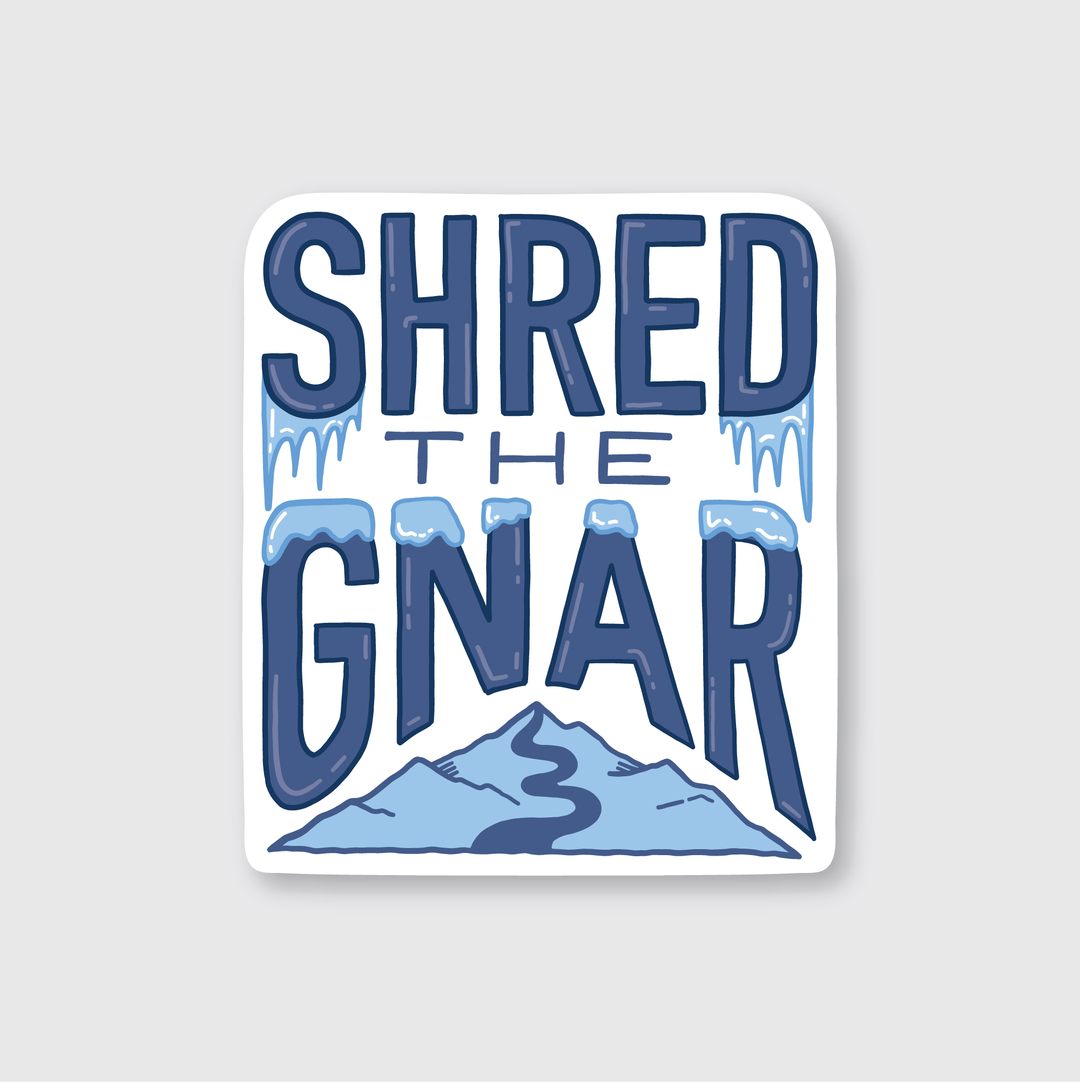 The "Shred The Gnar Sticker" features bold blue lettering accented with snow and icicles. It includes a mountain illustration with a winding path on a white background. This weather-resistant die-cut vinyl sticker is ideal for outdoor adventures.