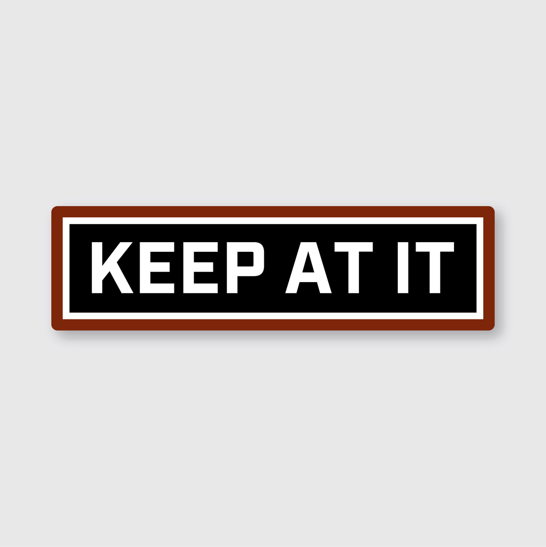 The "Keep At It Sticker" is a handcrafted vinyl sticker with a rectangular design. It features a dark background with bold white letters that spell out "KEEP AT IT," complemented by a striking red border. This motivational piece is crafted in Seattle and is perfect for adding inspiration to any space.