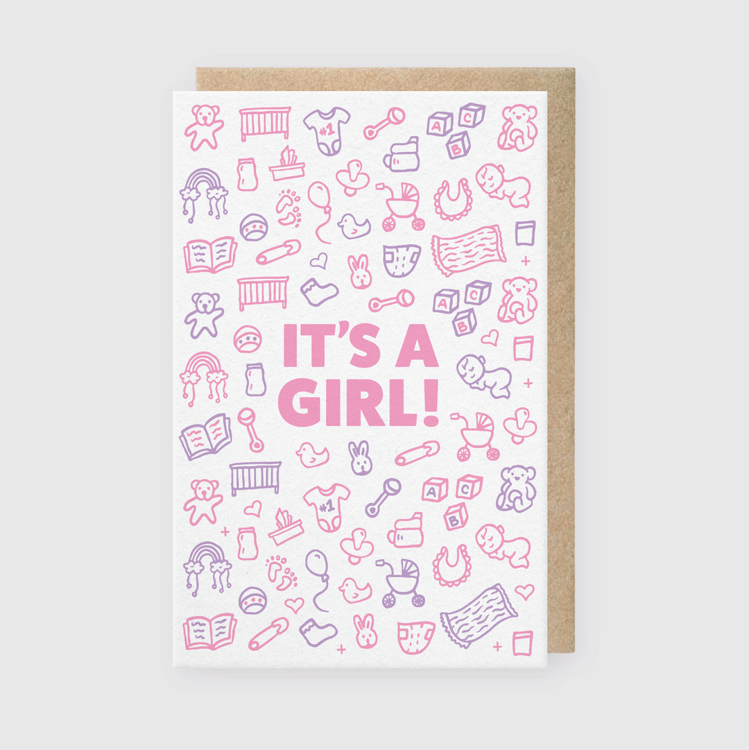 The "It's A Girl Greeting Card" features bold pink lettering surrounded by charming pink and purple doodles depicting baby items like bottles, teddy bears, rattles, and pacifiers. It's accompanied by a brown envelope and set against a light gray background.