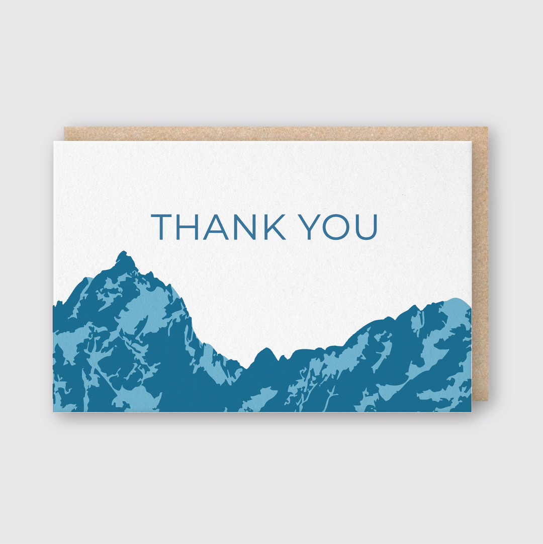 Introducing the Thank You Mountains Blue Greeting Card: This exquisite card features handcrafted blue mountain silhouettes near the bottom, with "THANK YOU" elegantly centered on a crisp white background. The premium letterpress design is beautifully paired with a classic brown envelope.