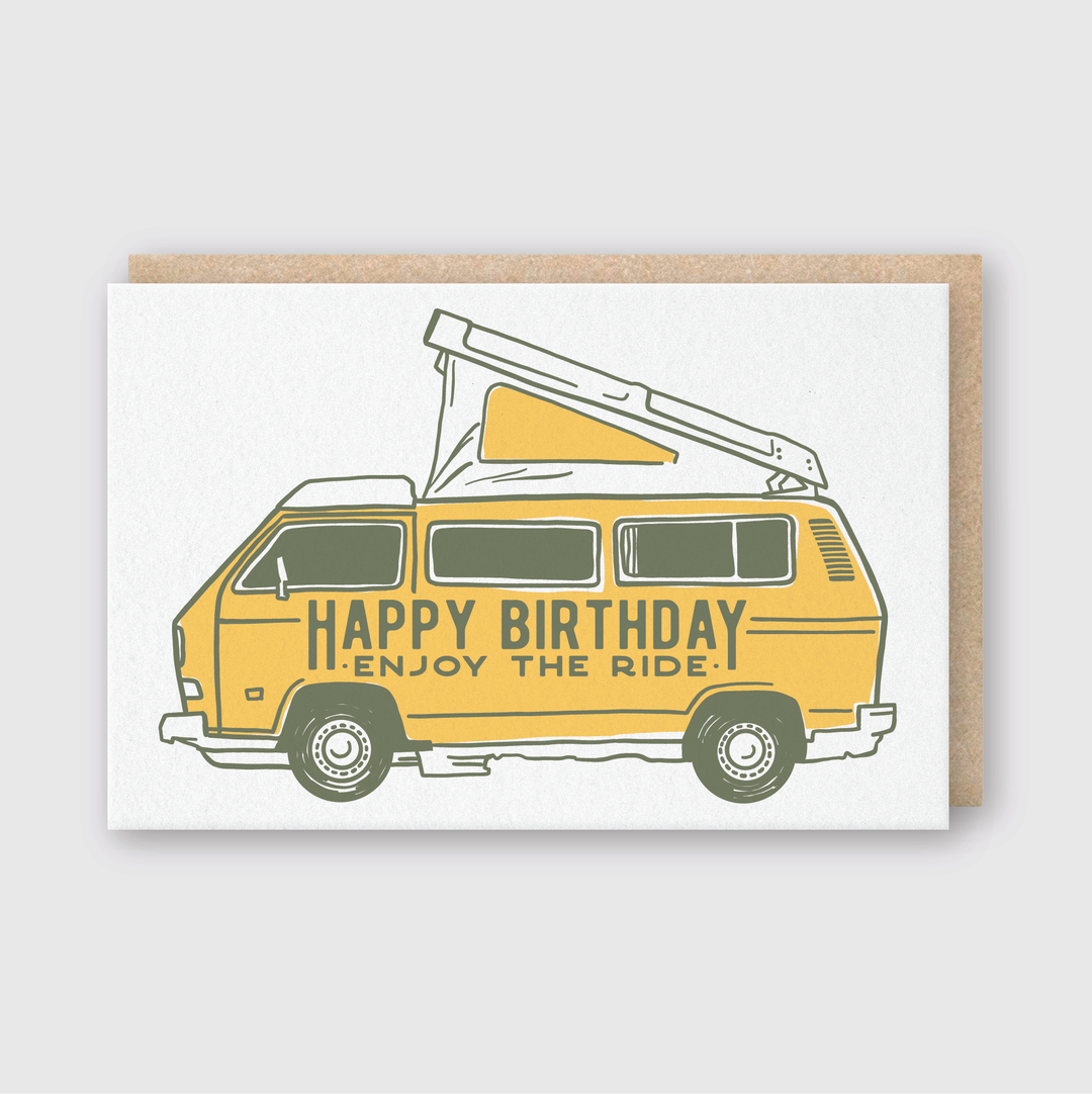 The Camper Van Birthday Greeting Card features an illustration of a yellow camper van with a pop-up roof, accompanied by the message "HAPPY BIRTHDAY, ENJOY THE RIDE" on its side. Crafted with expert letterpress techniques in Seattle, this card is presented on a light gray background and includes a partially visible brown envelope.
