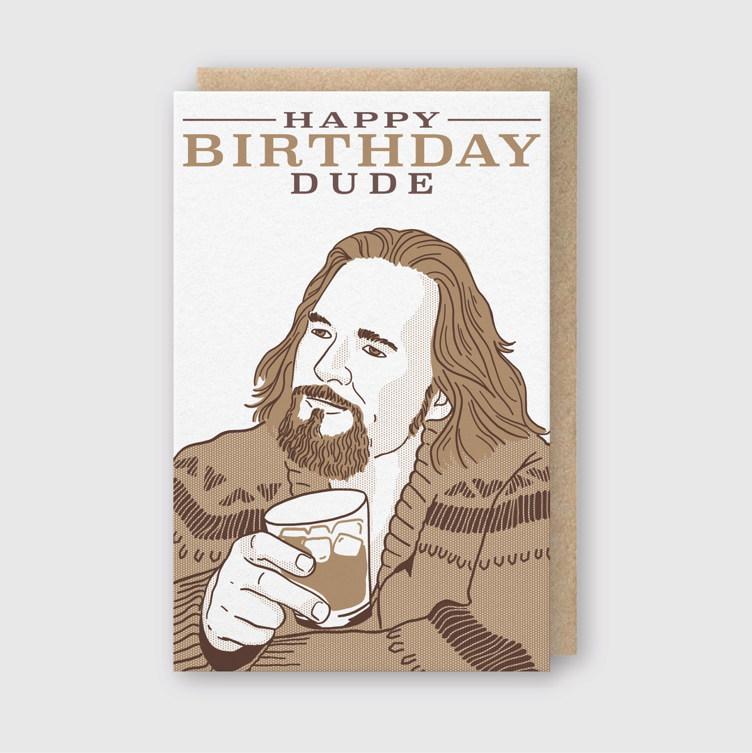 This handcrafted greeting card features an illustration of a bearded man with long hair holding a drink and wearing a patterned sweater. The "Happy Birthday Dude" message is prominently displayed in bold text, enhanced by an artisanal letterpress printing technique. The presentation is completed with a brown envelope.