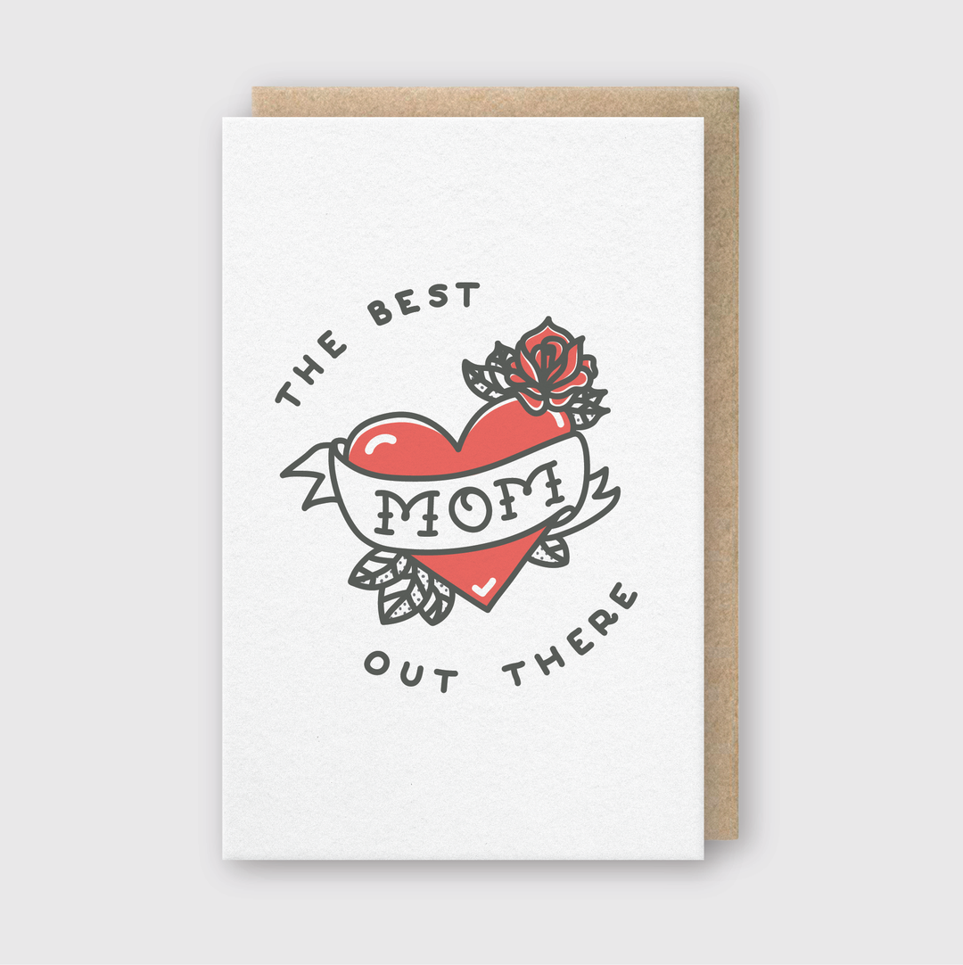 Introducing the Mom Heart Greeting Card, an exquisite handcrafted creation featuring a red heart and rose design centered with a "MOM" banner. The elegant letterpress card includes the phrases "THE BEST" above and "OUT THERE" below, showcased beautifully on a stylish brown envelope against a light gray background. This card is ideal for honoring the incredible mom in your life!