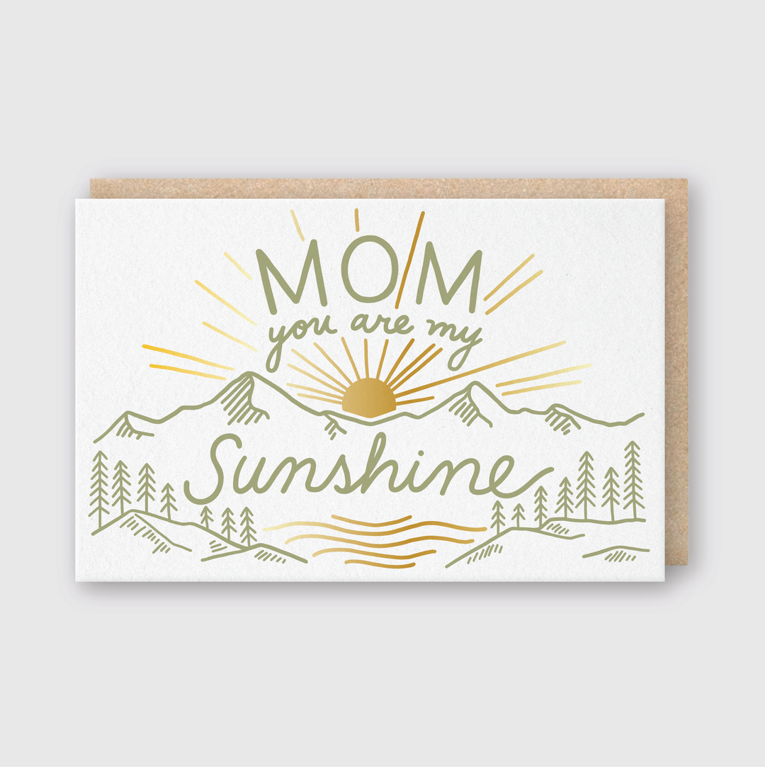 Introducing the "Mom You Are My Sunshine" Greeting Card. This exquisite, handcrafted card showcases a hand-drawn design of a sun rising over mountains and trees, accompanied by the heartfelt message "Mom, you are my Sunshine" in vibrant green and yellow hues. Perfect for conveying warmth and appreciation.