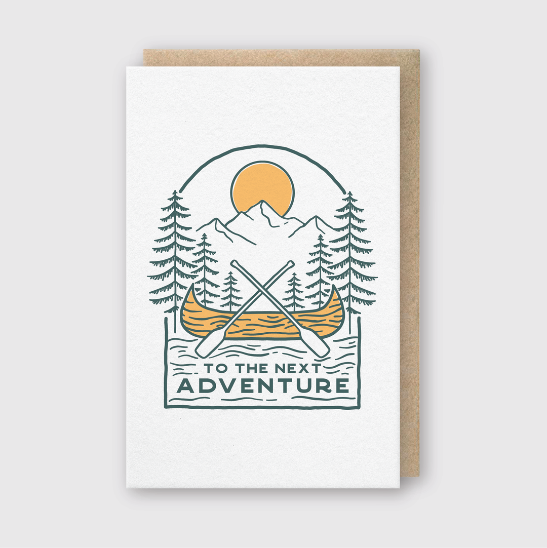 The "To The Next Adventure Greeting Card" offers a beautifully crafted letterpress design. It highlights a canoe with crossed paddles on a serene lake, surrounded by majestic pine trees and tall mountains under a setting sun. The card carries the message, "To the next adventure.