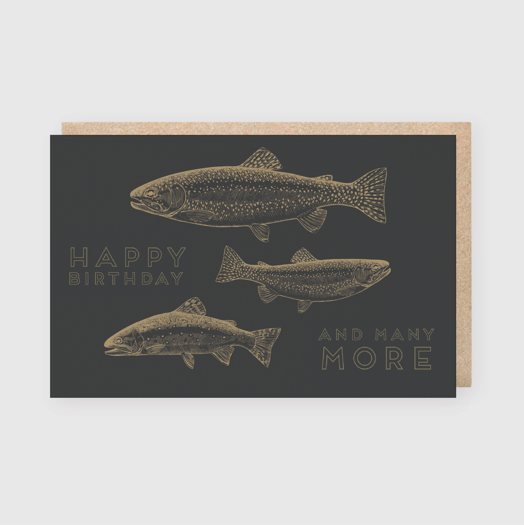 Introducing the Happy Birthday & Many More Greeting Card, which showcases three illustrated fish against a dark background. Crafted using letterpress printing, this card carries the inscription "Happy Birthday and Many More" and is handmade in Seattle. It comes with a brown paper envelope that is partially visible behind it.