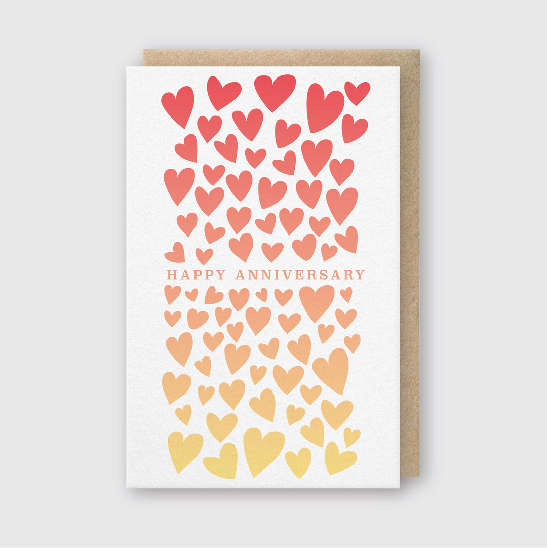 The Happy Anniversary Split Greeting Card features the words "Happy Anniversary" surrounded by a cascade of hearts transitioning from red to yellow on a light gray background. Handcrafted in Seattle, it beautifully captures the essence of celebration and love.