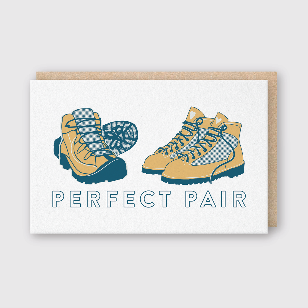 The Perfect Pair Hikers Greeting Card is designed for outdoor lovers, featuring two hiking boots with contrasting accents above the phrase "Perfect Pair." The card has a simple gray background and comes with a brown paper insert. It's handcrafted in Seattle.