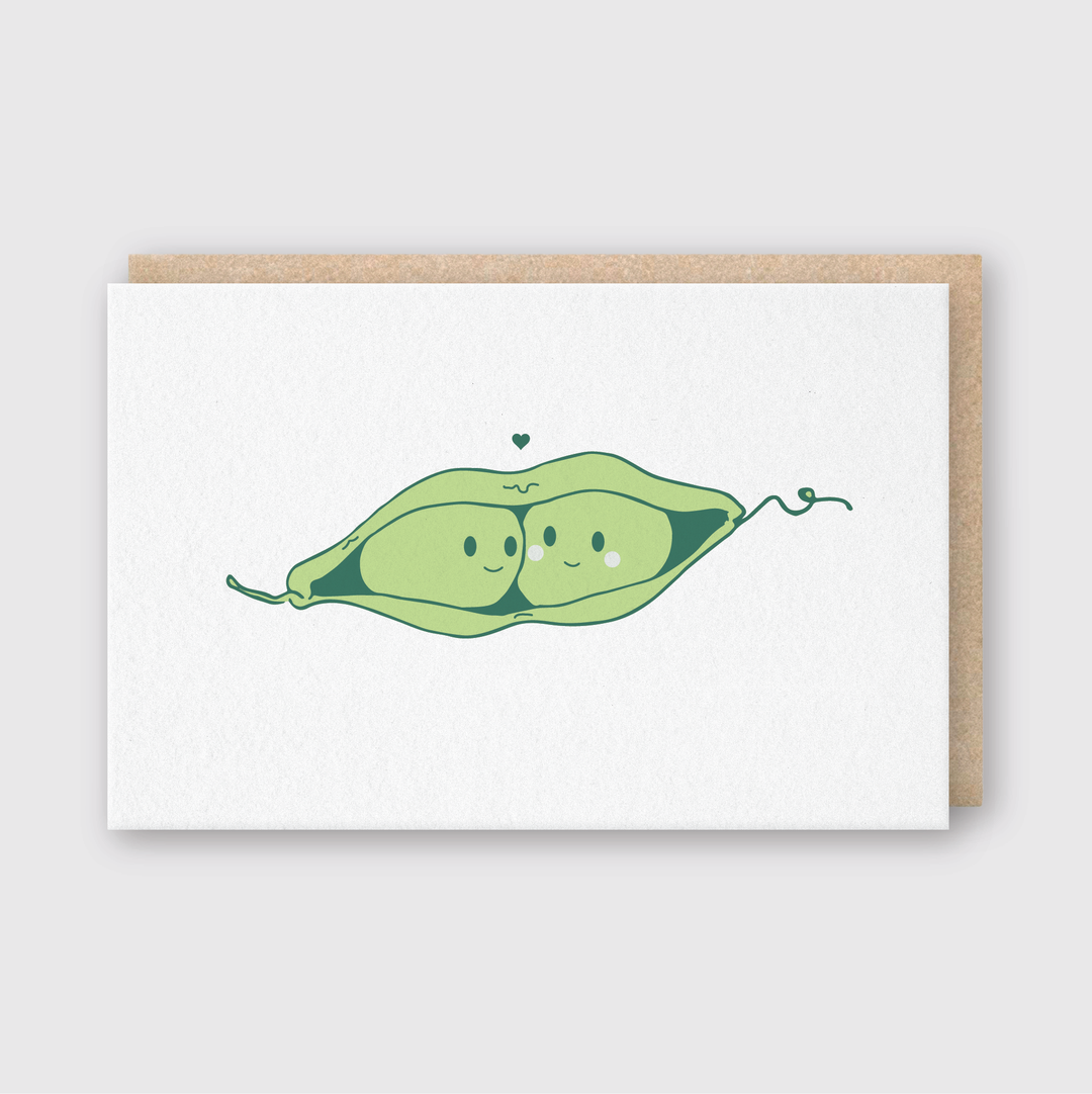 The Lover Peas Greeting Card showcases an illustration of two cheerful peas nestled in an open pod, with a small heart above them. Set against a plain white background, the card includes a partially visible brown paper envelope. This card is thoughtfully handcrafted using environmentally friendly soy-based inks.
