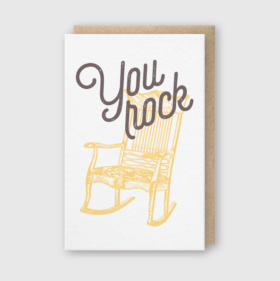 The greeting card features a simple yet vibrant design, with a yellow rocking chair illustration and the phrase "You rock" written in bold, cursive letters at the top. It stands out against a plain white background and comes with a light brown envelope that can be seen partly from behind.