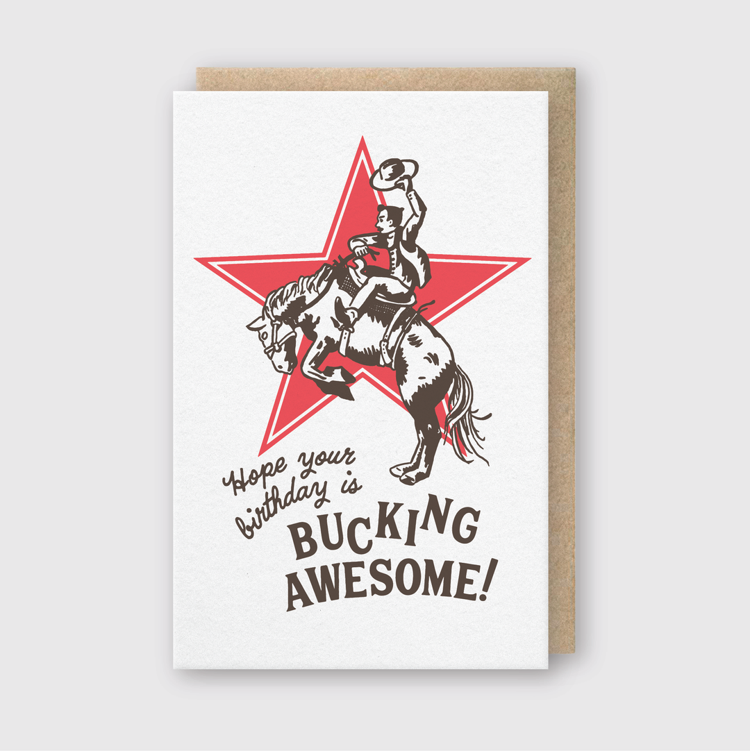 The Bucking Birthday Greeting Card features a cowboy on a bucking horse with a shining red star in the background. It includes the message, "Hope your birthday is BUCKING AWESOME!" and comes with a brown envelope, showcasing handcrafted details.