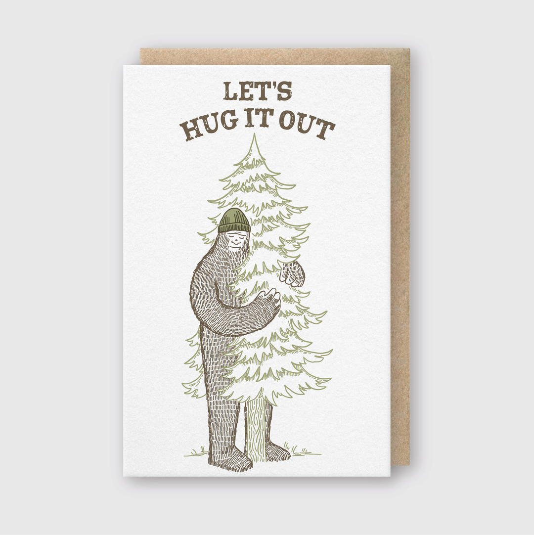 The Hug It Out Greeting Card features a Sasquatch in a beanie embracing a pine tree, along with the phrase "LET'S HUG IT OUT." Handmade in Seattle, it's paired with a recycled kraft envelope and has a textured brown border.
