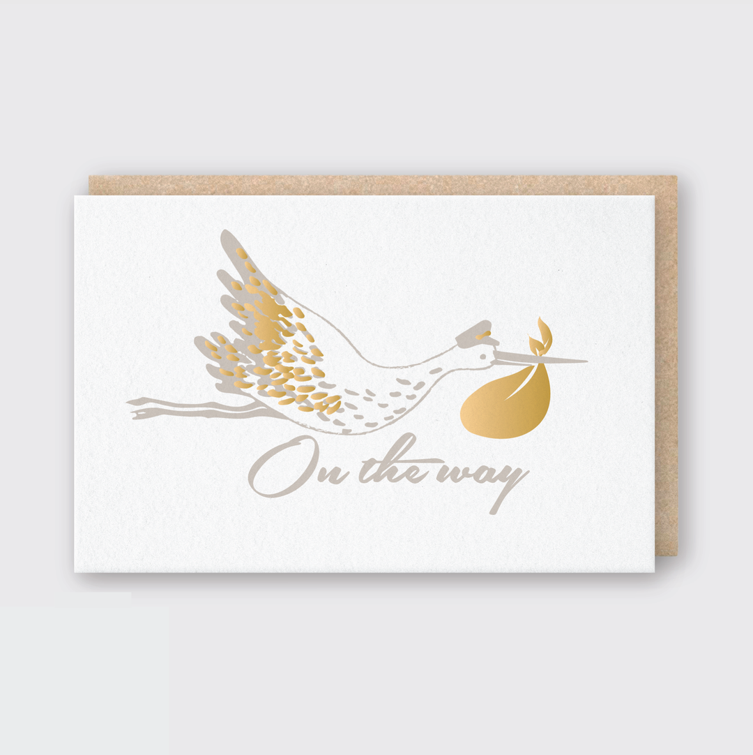 The Baby On The Way Greeting Card showcases an illustrated stork carrying a golden bundle, with the phrase "On the way" elegantly inscribed below. It is accompanied by a classic brown envelope.
