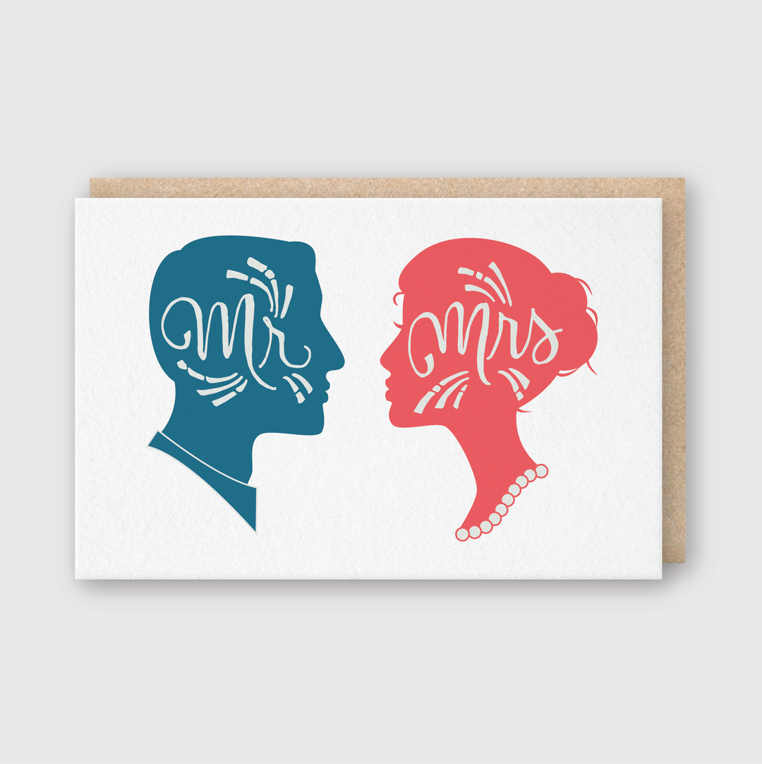 This handcrafted Mr. Mrs. Silhouette Greeting Card features a blue silhouette of a man's face with "Mr" and a pink silhouette of a woman's face with "Mrs," accented by a pearl necklace. It is elegantly presented on a white card paired with a beige envelope, crafted in Seattle.