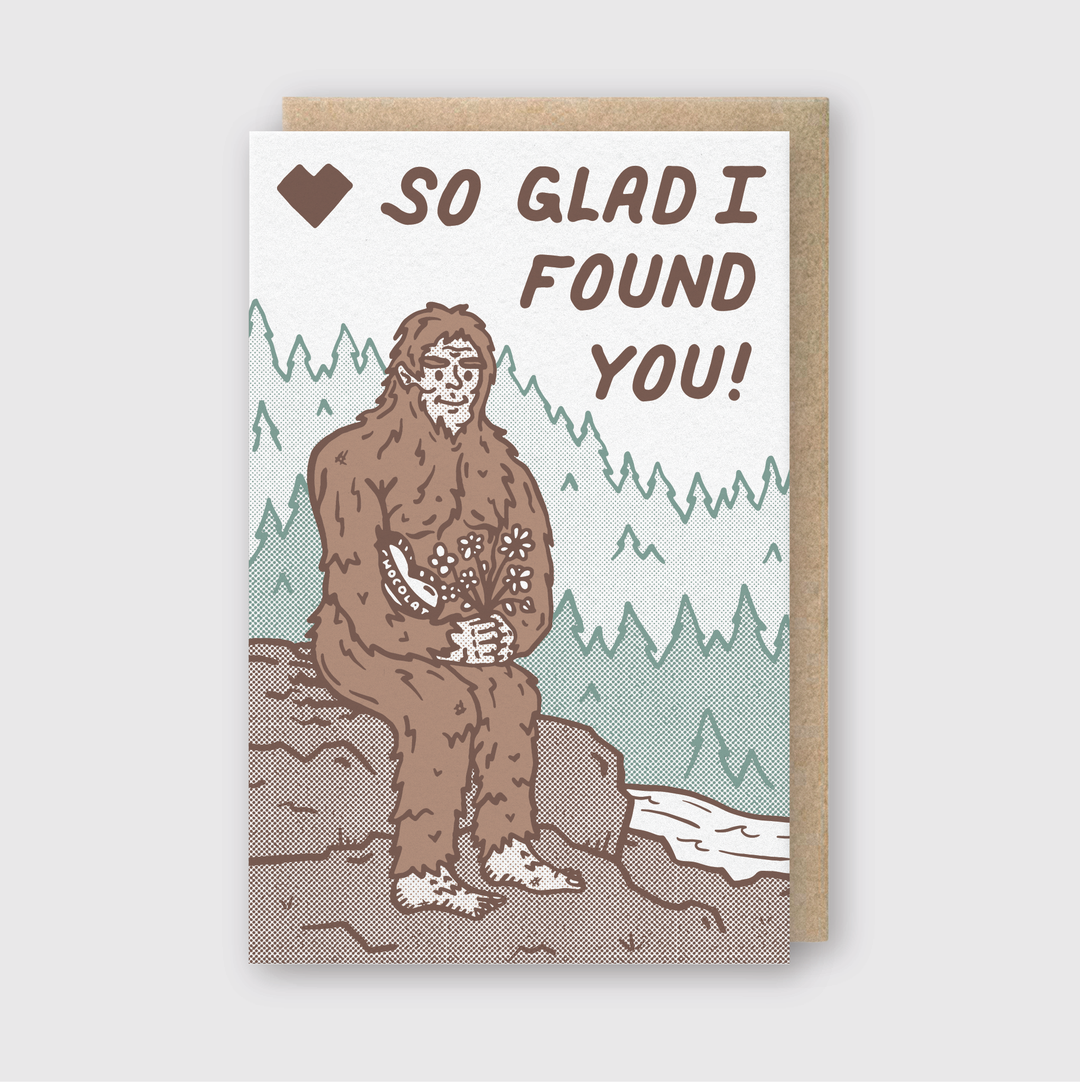 The greeting card features a whimsical illustration of Bigfoot sitting on a rock, holding flowers. Above Bigfoot, the words "So glad I found you!" appear alongside a heart icon. The background is filled with trees, and it comes with a recycled Kraft envelope.