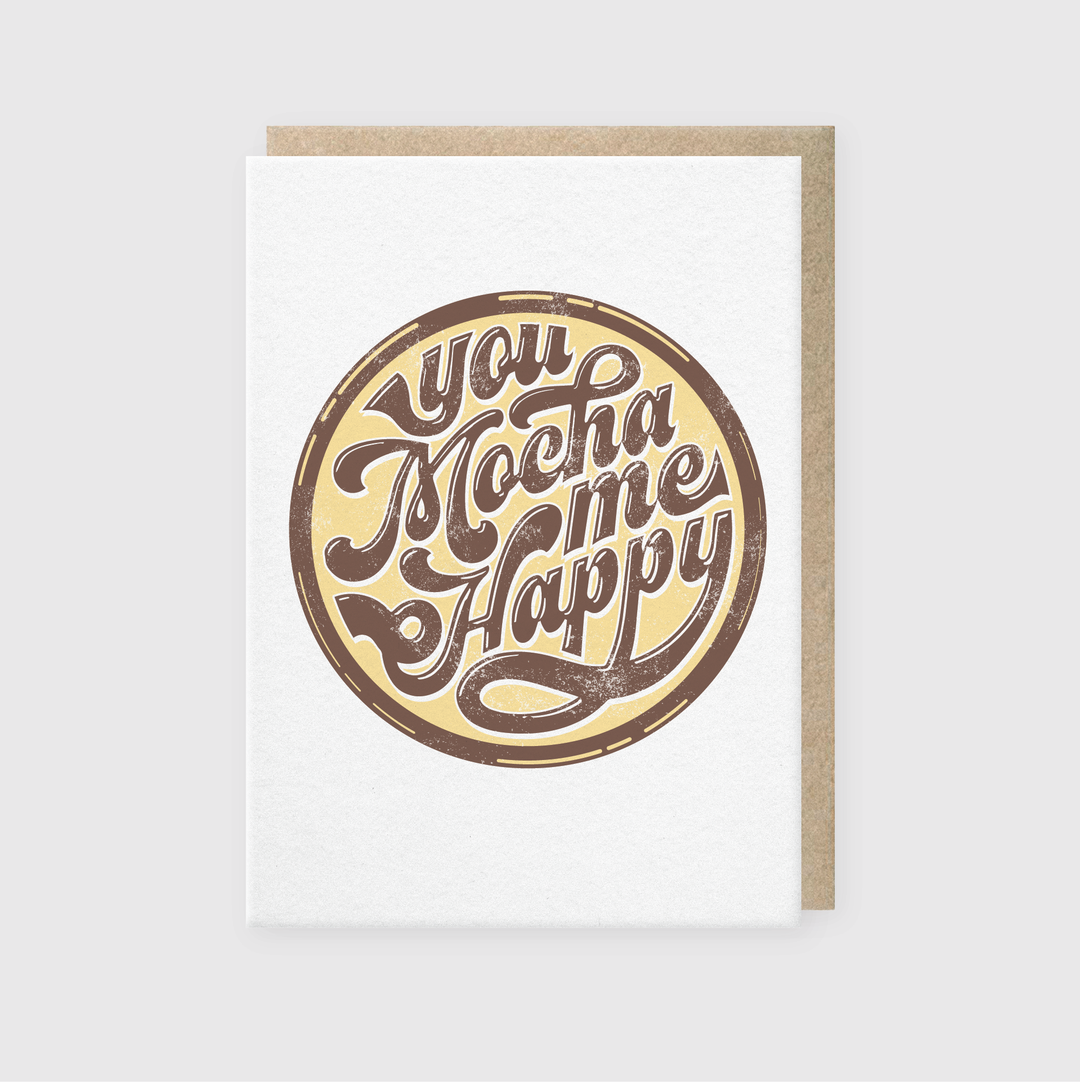 Introducing the "You Mocha Me Happy Greeting Card," a delightful option for coffee enthusiasts. This card showcases a circular design with warm brown and cream tones on a gray background, giving it an inviting and cozy feel. When partially opened, it reveals a luxurious brown interior, enhancing its charm and appeal.