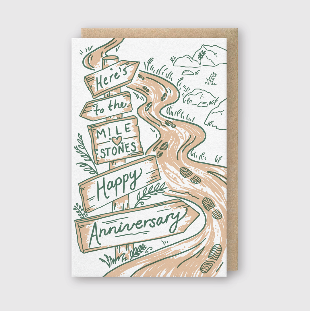The Milestones Anniversary Greeting Card features a winding path adorned with hand-printed signposts bearing the messages "Here's to the Milestones" and "Happy Anniversary." The card is set against a nature-inspired backdrop with rocks and plants, crafted on FSC-certified paper for an eco-friendly appeal.