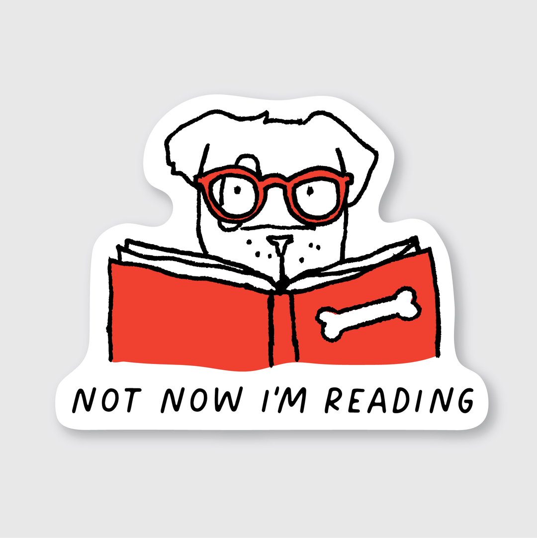 The "Not Now I'm Reading Sticker" features a charming design of a dog wearing red glasses and reading a red book with a bone on the cover. Below, the text reads, "Not Now I'm Reading." This die-cut vinyl sticker is created to be weather-resistant, making it perfect for showcasing your love for reading in any weather.