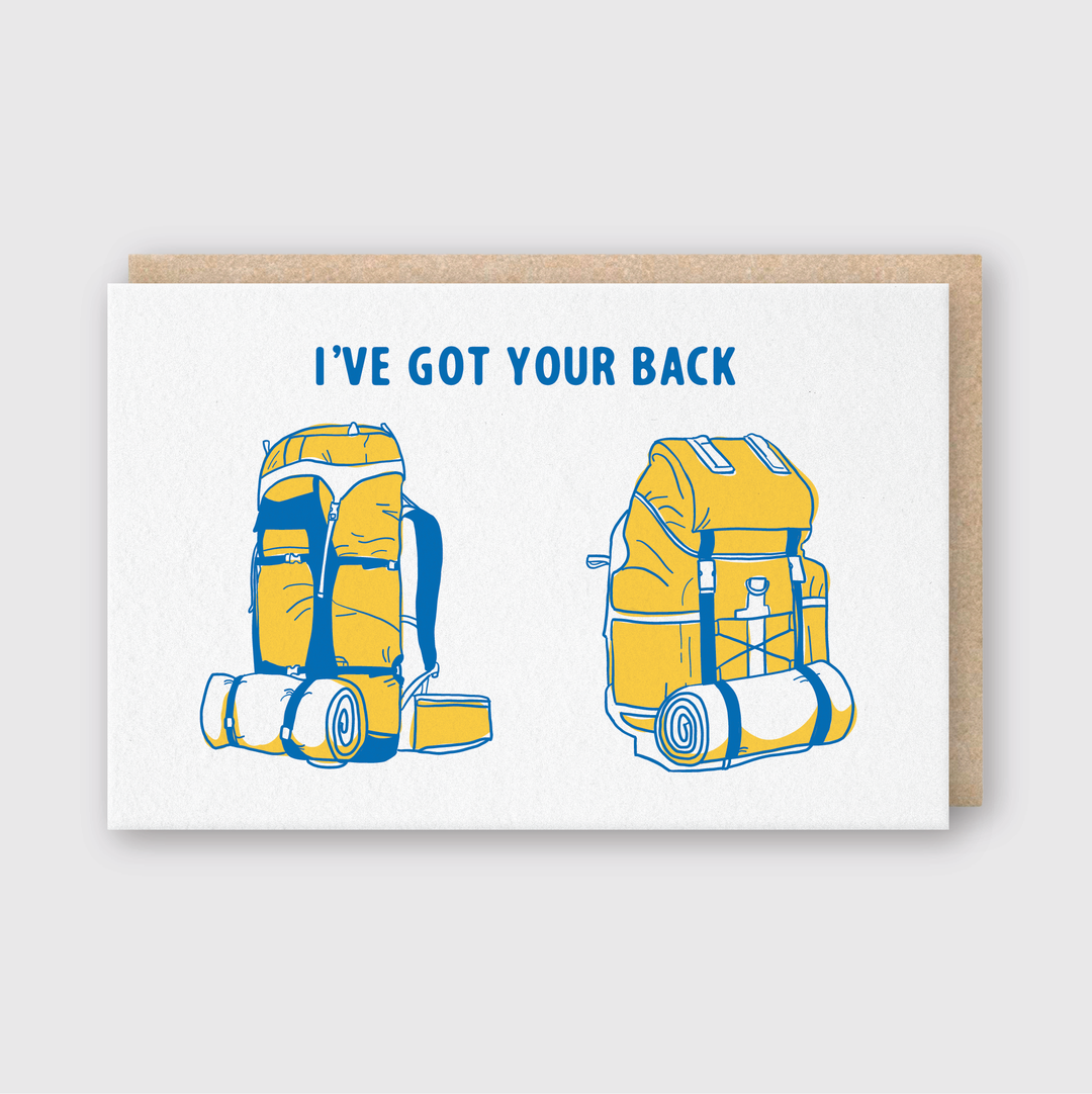 The "I've Got Your Back Greeting Card" showcases an illustration of two backpacks, one yellow and one blue, depicted from both front and back views. This card is handcrafted in Seattle, printed on FSC Certified Paper, and comes with a brown envelope. The presentation features a gray background.