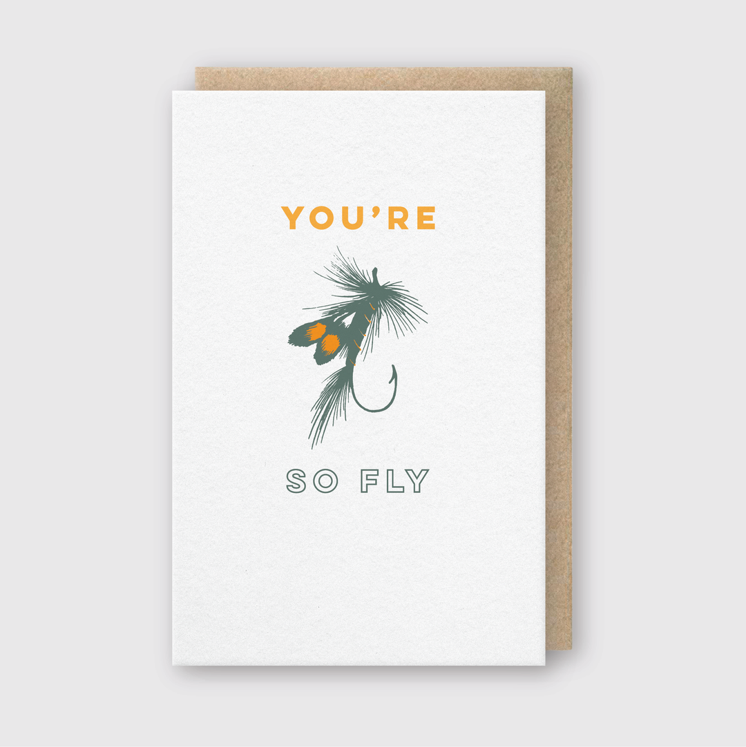 The "You’re So Fly" Hook Greeting Card showcases a fishing fly lure design with the words "You're So Fly" in vibrant orange and green. This card is expertly handcrafted in Seattle and uses FSC Certified Paper. It comes with a brown envelope against a light gray background.