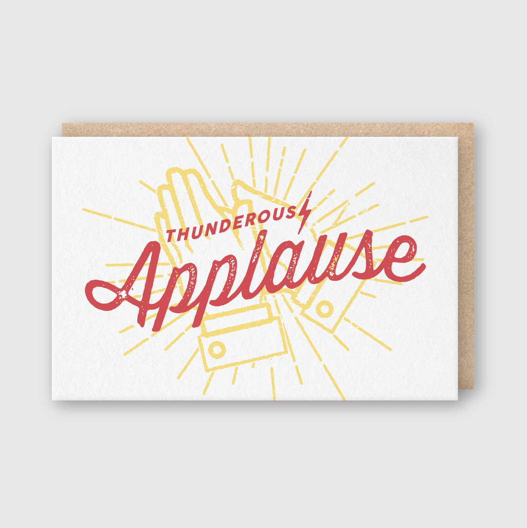 The Thunderous Applause Greeting Card features a design with two clapping hands outlined in yellow. The phrase "Thunderous Applause" is elegantly scripted in bold red cursive over the hands, all set against a white background. It comes with a subtly peeking beige envelope.