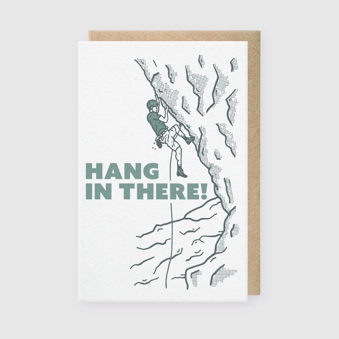 A handcrafted greeting card featuring a minimalist illustration of a rock climber in helmet and harness scaling a rocky surface, with the encouraging message "Hang In There!" This card offers an uplifting sentiment against a neutral background.