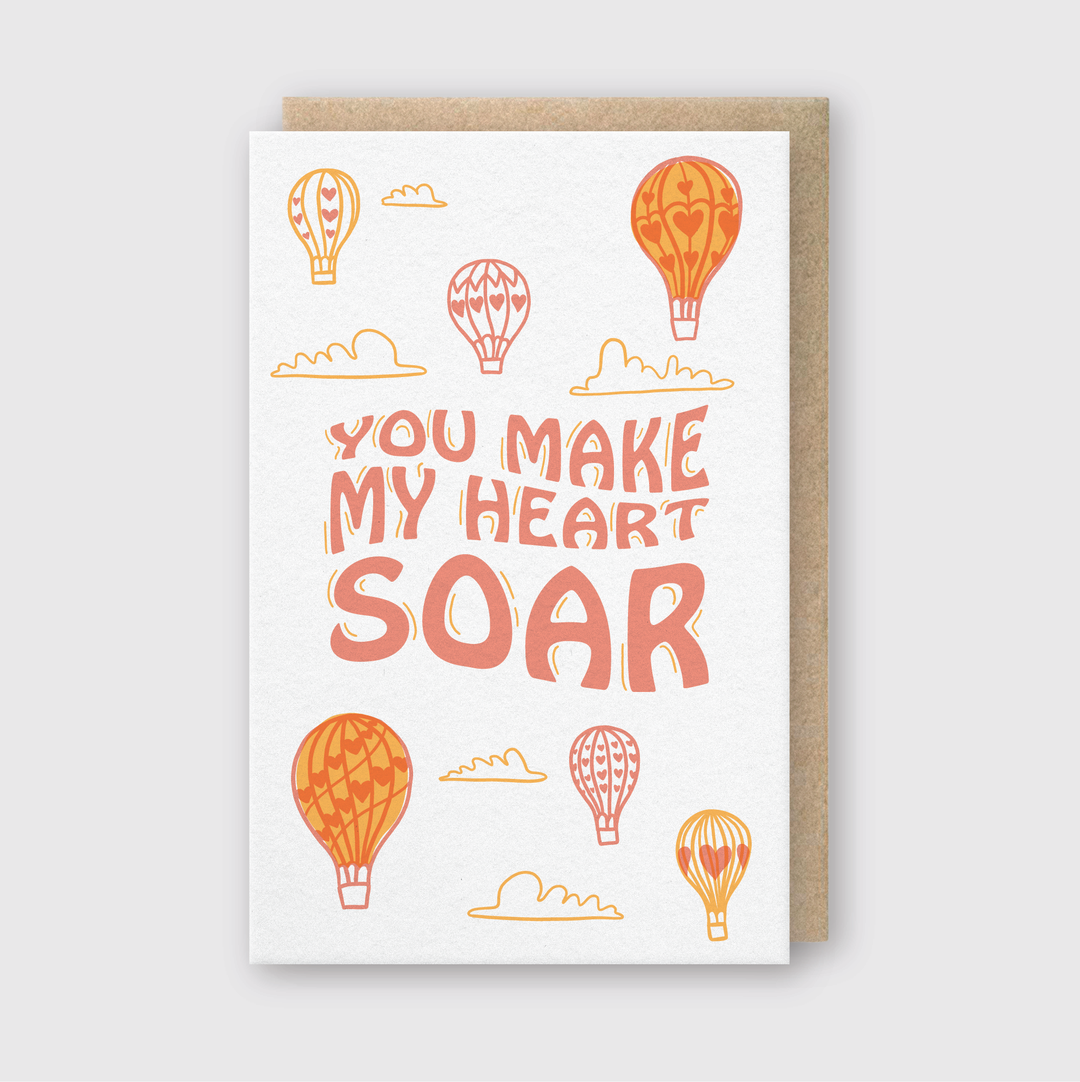The "You Make My Heart Soar" Greeting Card features beautiful illustrations of orange and red hot air balloons with clouds, crafted on FSC Certified Paper, and is showcased on a brown envelope.