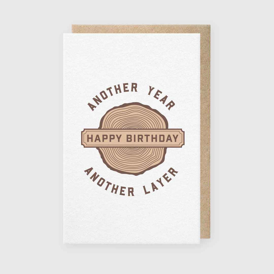 The "Another Year, Another Layer Greeting Card" features a unique tree ring design at its center, crafted with letterpress techniques. Inside the ring, the words "Happy Birthday" are elegantly displayed, surrounded by the phrase "Another Year Another Layer" in brown letters. It includes a recycled Kraft envelope for an eco-friendly touch and is set against a light gray background for added sophistication.