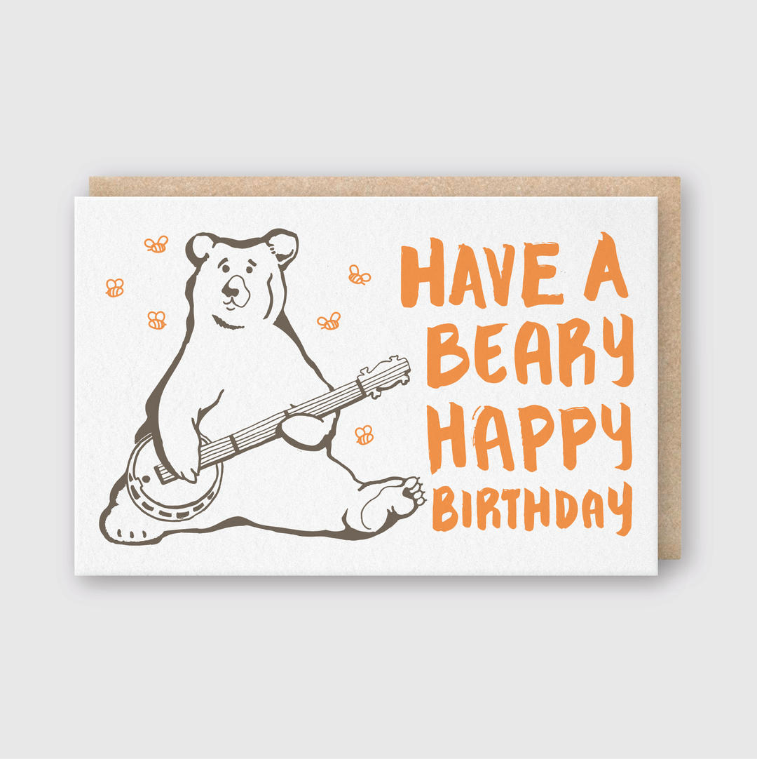 The Beary Happy Birthday Greeting Card features an adorable bear playing a banjo. Crafted through letterpress printing, it displays the message "Have a Beary Happy Birthday" in bold orange letters, complemented by lovely orange flowers for a cozy accent.