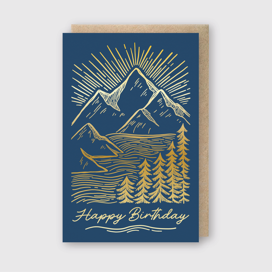 The Birthday Mountainscape Greeting Card features a blue background with gold letterpress details, showcasing mountains, trees, and sun rays. Made from FSC-certified paper, the card includes an elegant script at the bottom that reads "Happy Birthday.