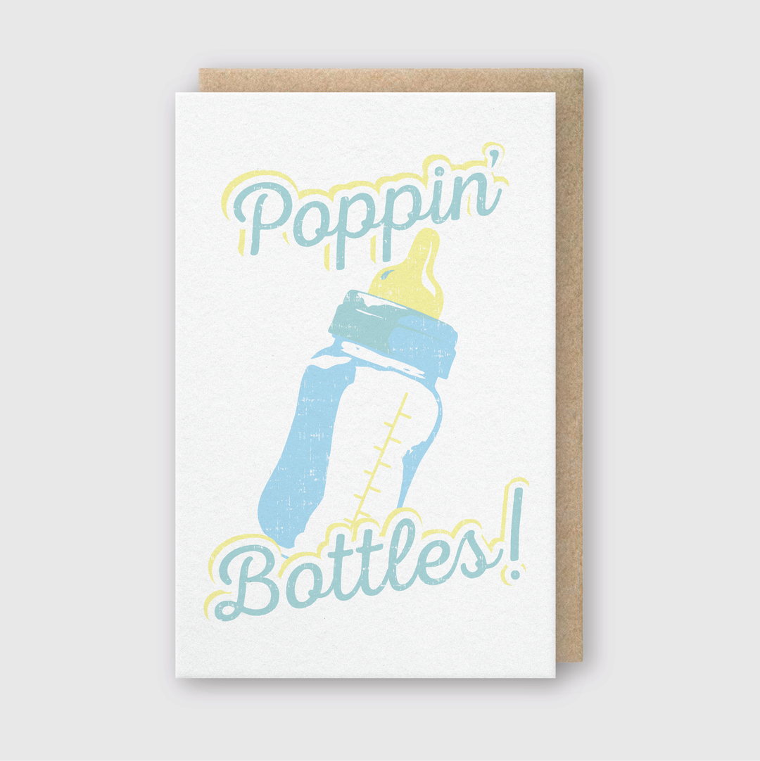 The "Poppin Bottles Greeting Card" features a charming letterpress design with a baby bottle graphic and the phrase "Poppin' Bottles!" in playful blue and yellow font on a clean white background. The card is attractively set against a light gray backdrop, making it perfect for celebrating new arrivals.