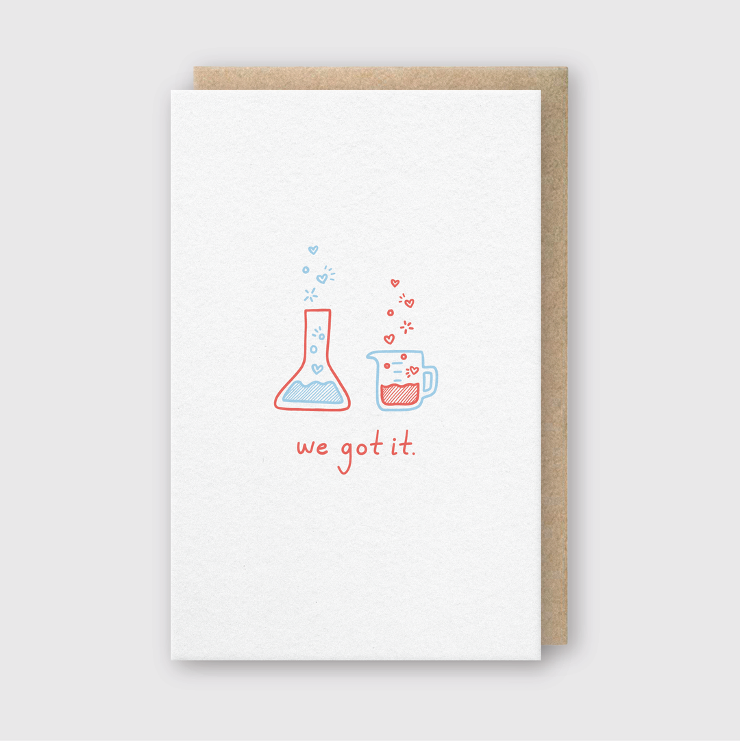 The We Got It Chemistry Greeting Card features delightful illustrations of a blue chemistry flask and a red coffee mug adorned with heart designs. A bold red caption saying "we got it" adds an extra layer of style. This handcrafted card is beautifully paired with a recycled kraft envelope, all set against a subtle light gray background.