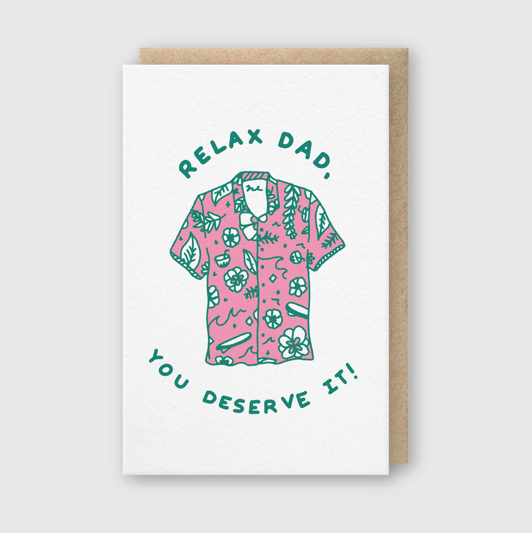 The "Relax Dad" Greeting Card features a pink Hawaiian shirt with green tropical patterns as its centerpiece. The card displays the words "Relax Dad" above the shirt and "You deserve it!" below. Crafted by hand, it comes against a plain backdrop with a brown envelope included.