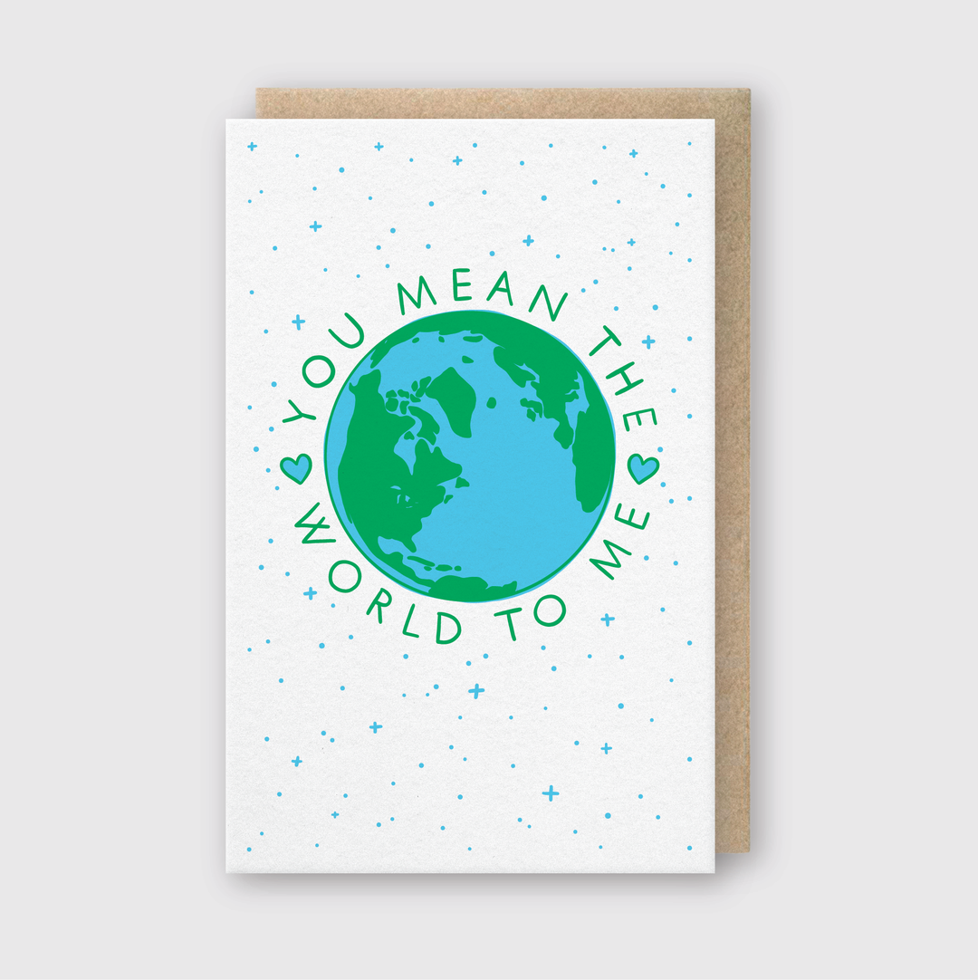 The "You Mean The World To Me" greeting card features a charming illustration of Earth in blue and green, surrounded by stars. It is hand printed in Seattle on FSC Certified Paper and adorned with small heart symbols. The card comes with a matching brown envelope, enhancing its delightful presentation.