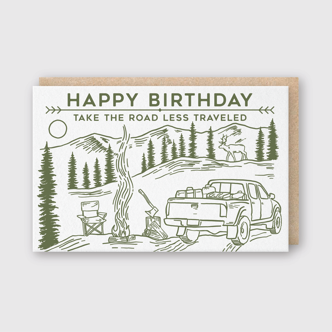 The Happy Birthday Road Less Traveled Truck Greeting Card by Pike Street Press depicts a charming picnic scene with a pickup truck, campfire, and chairs amidst stunning mountains and trees. A curious deer looks on as the card displays the message "HAPPY BIRTHDAY" along with "TAKE THE ROAD LESS TRAVELED.
