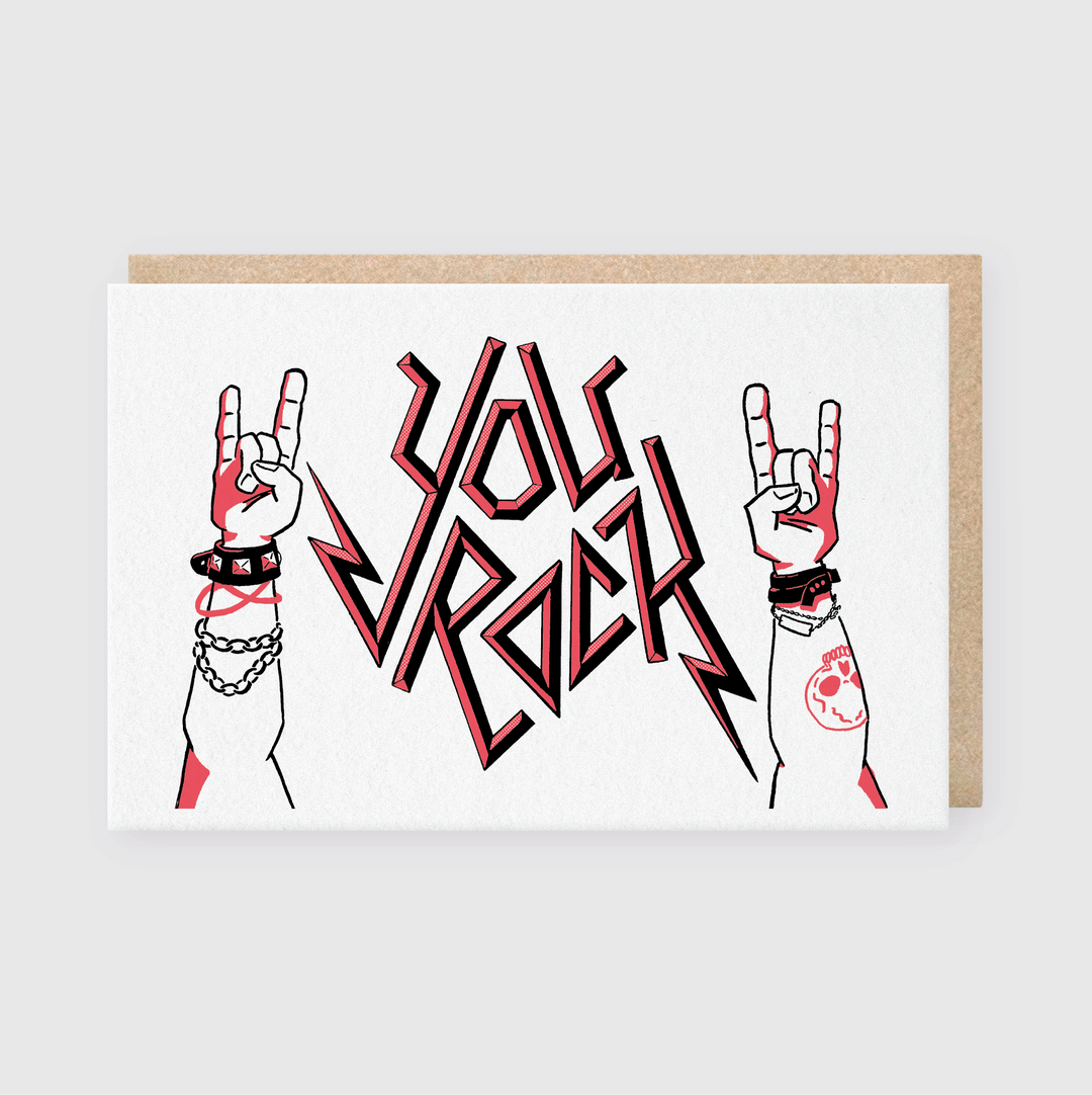 The You Rock Greeting Card is a handcrafted creation featuring a recycled Kraft envelope. It showcases two hands making the "rock on" sign, with bold angular letters declaring "You Rock" between them. One wrist is embellished with chains and bracelets, and the design boasts a striking combination of black, red, and white colors crafted using letterpress techniques.