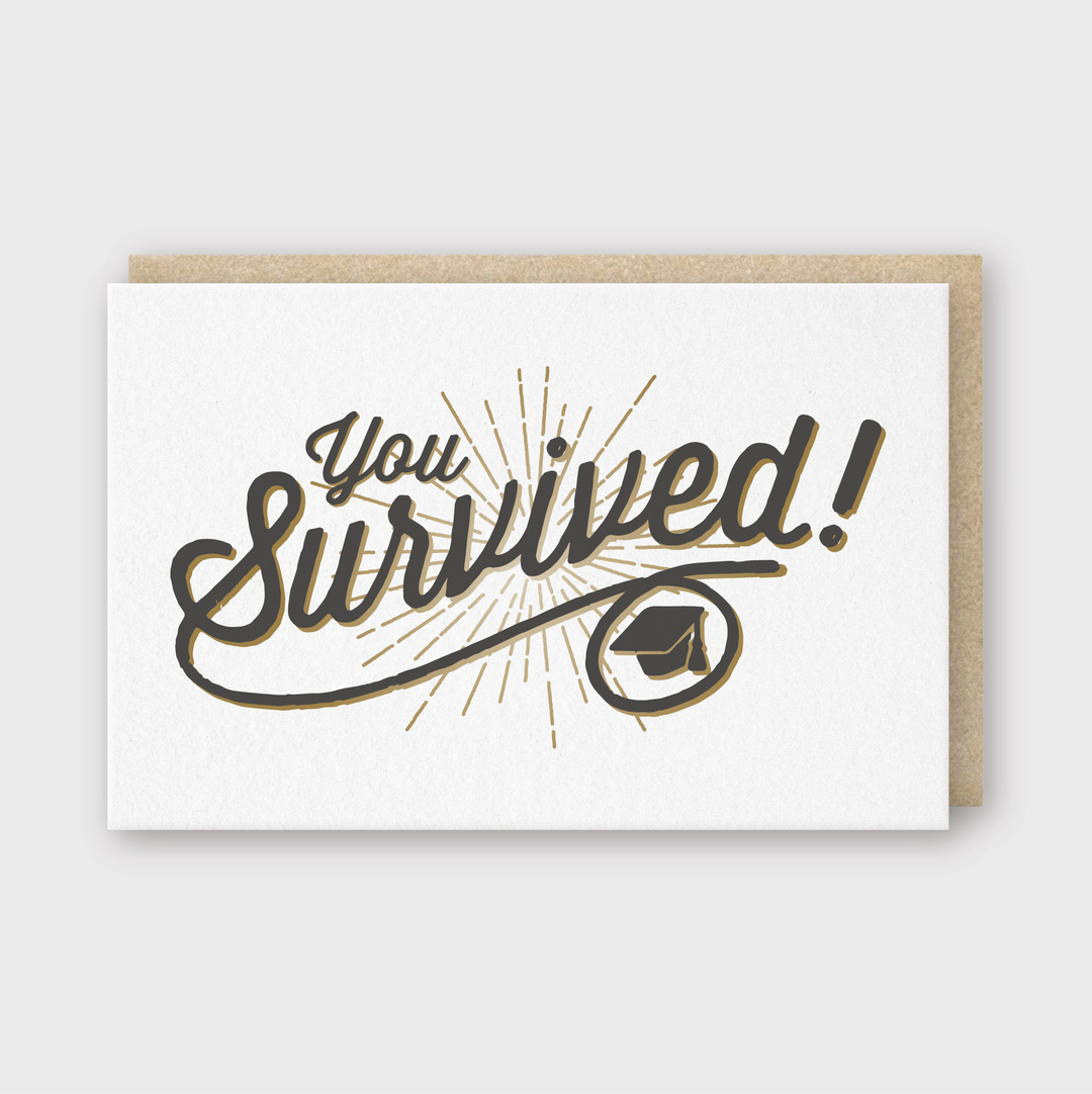 The "You Survived Greeting Card" showcases the phrase "You Survived!" in a bold, stylized font along with an icon of a graduation cap accented with radiant lines, adding a celebratory touch. This card is handcrafted in Seattle and printed on FSC Certified Paper, making it an ideal choice for commemorating any significant achievement. Its design is simple yet impactful.