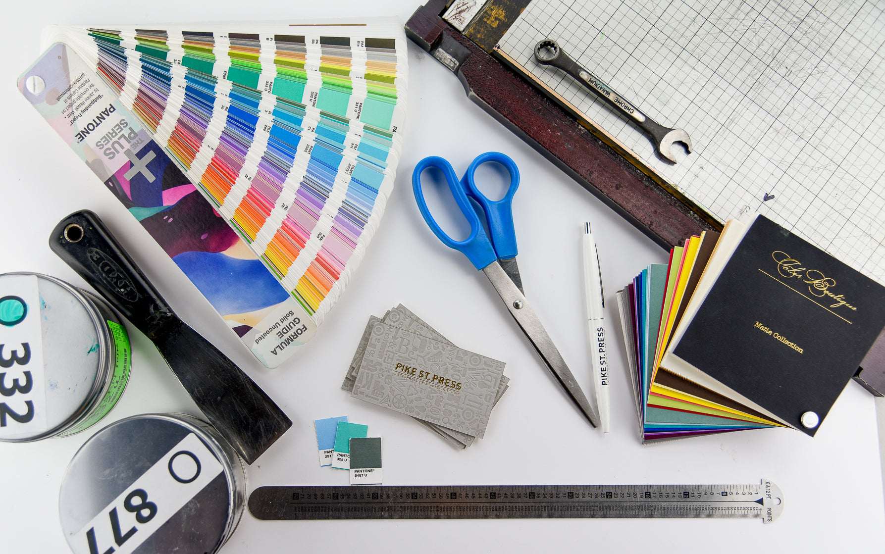 A creative workspace is laid out on a white surface, featuring an assortment of design tools. Among the items are a color swatch, scissors, a paper cutter, and a ruler. There’s also a wrench and several paint cans present. The scene is complemented by business cards and various color samples, enhancing the atmosphere of creativity and design exploration.