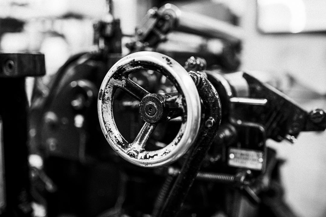 Close-up of a vintage mechanical machine, featuring a worn metal wheel with a five-spoke design. The machine, shown in black and white, has a rustic, industrial appearance with intricate gears and levers in the background.