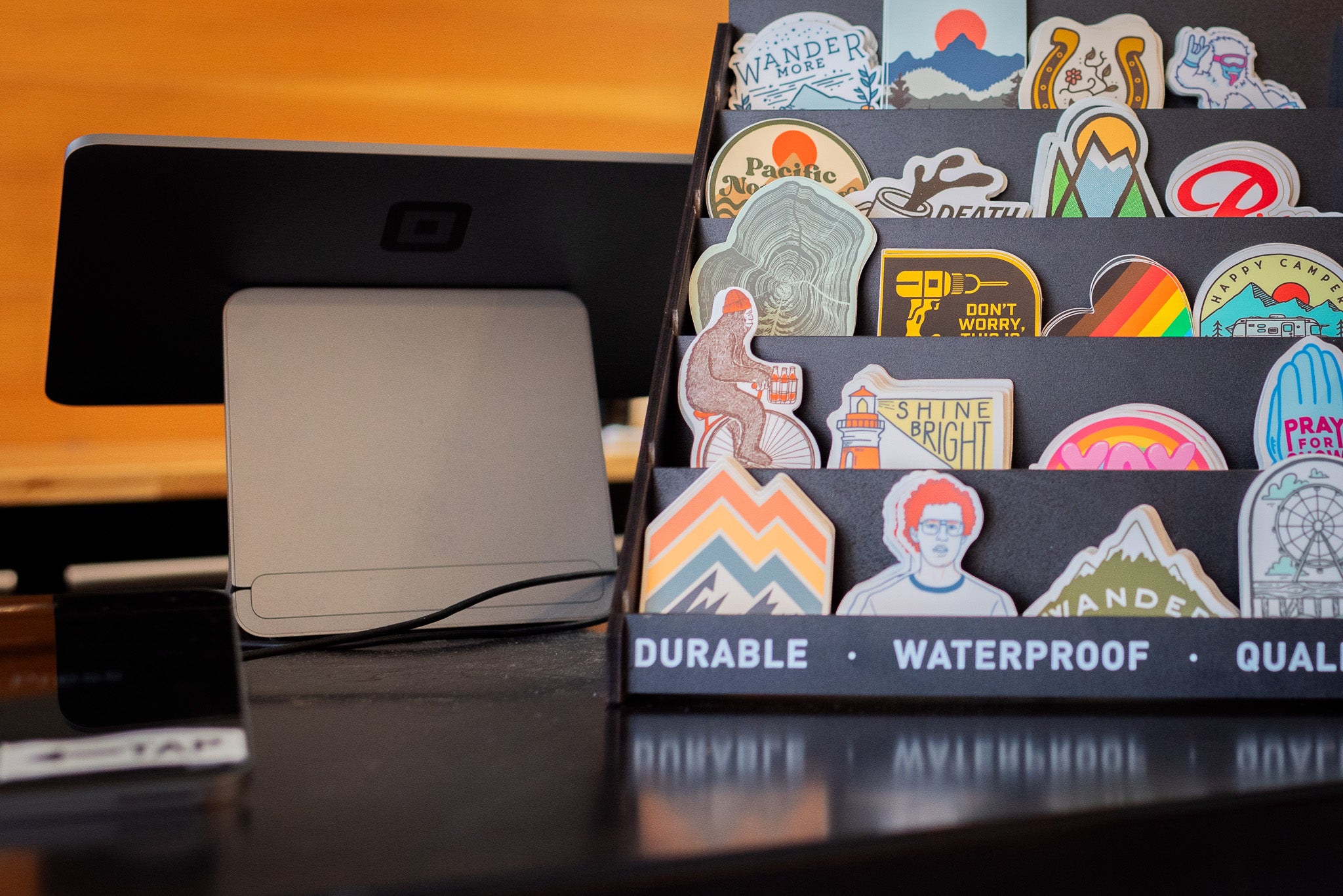 The scene features a vibrant and creative setup. On the right, there's a black stand adorned with an assortment of colorful stickers showcasing designs such as mountains, animals, and inspirational quotes, adding a lively touch to the display. To the left is a digital point-of-sale terminal positioned on the countertop, likely used for processing transactions efficiently. The combination of these elements suggests a modern and artistic retail environment.