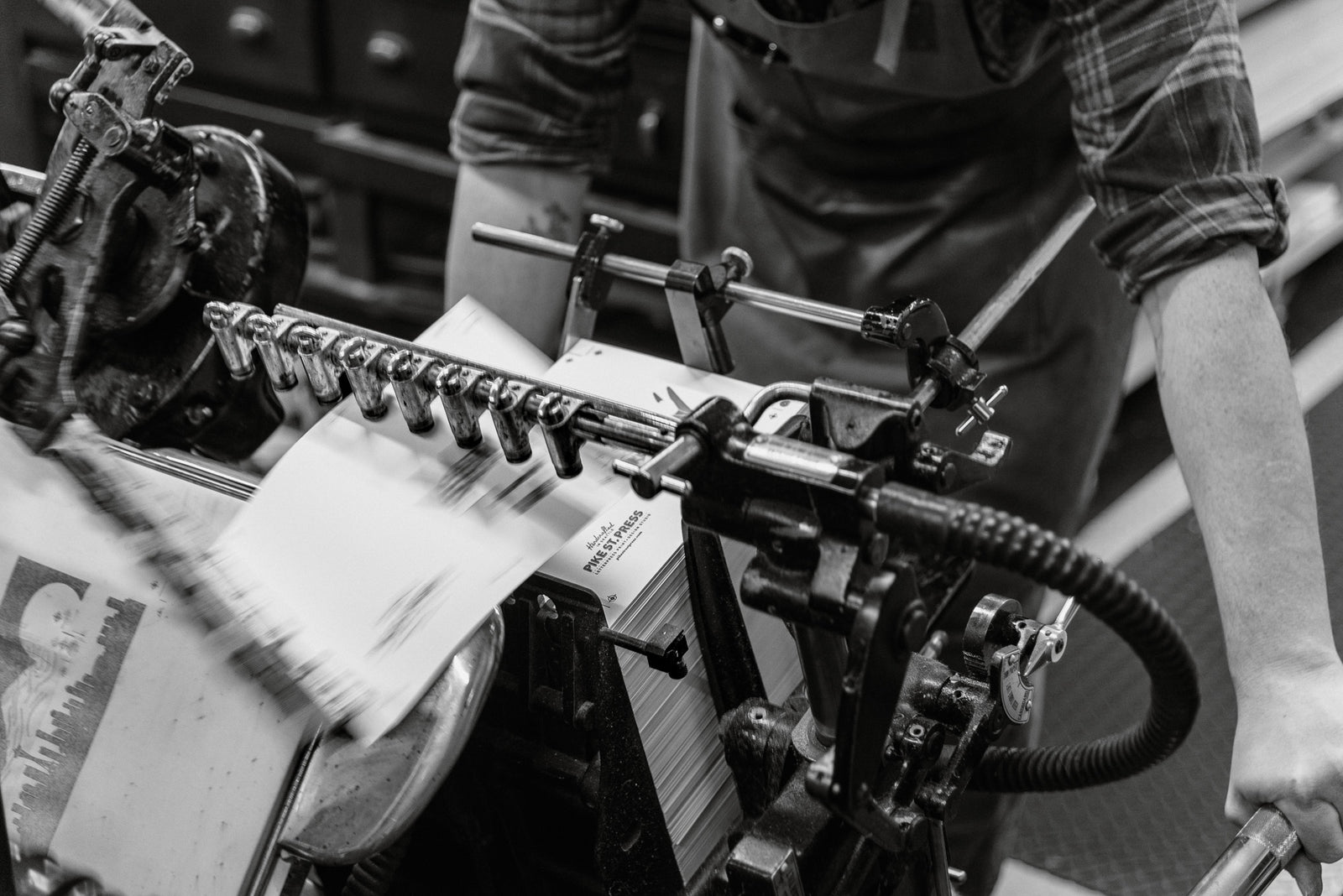 This black and white photo captures an individual diligently operating a vintage printing press. The machine is in dynamic motion, with sheets of paper collecting neatly on one side. The operator, dressed in an apron, displays focused attention on the intricate workings of the press. The image evokes a sense of craftsmanship and dedication to the art of printing from a bygone era.