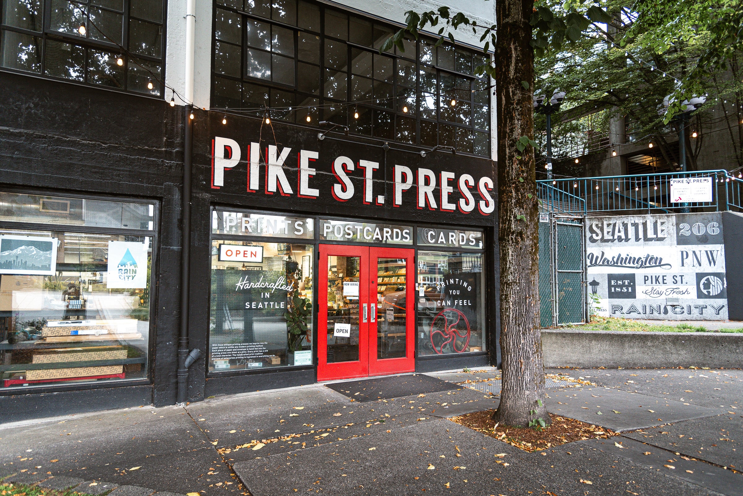 A storefront features red double doors with signs indicating it's open and offering prints, postcards, and cards. The exterior includes trees adorned with string lights, and the sidewalk is scattered with fallen leaves.