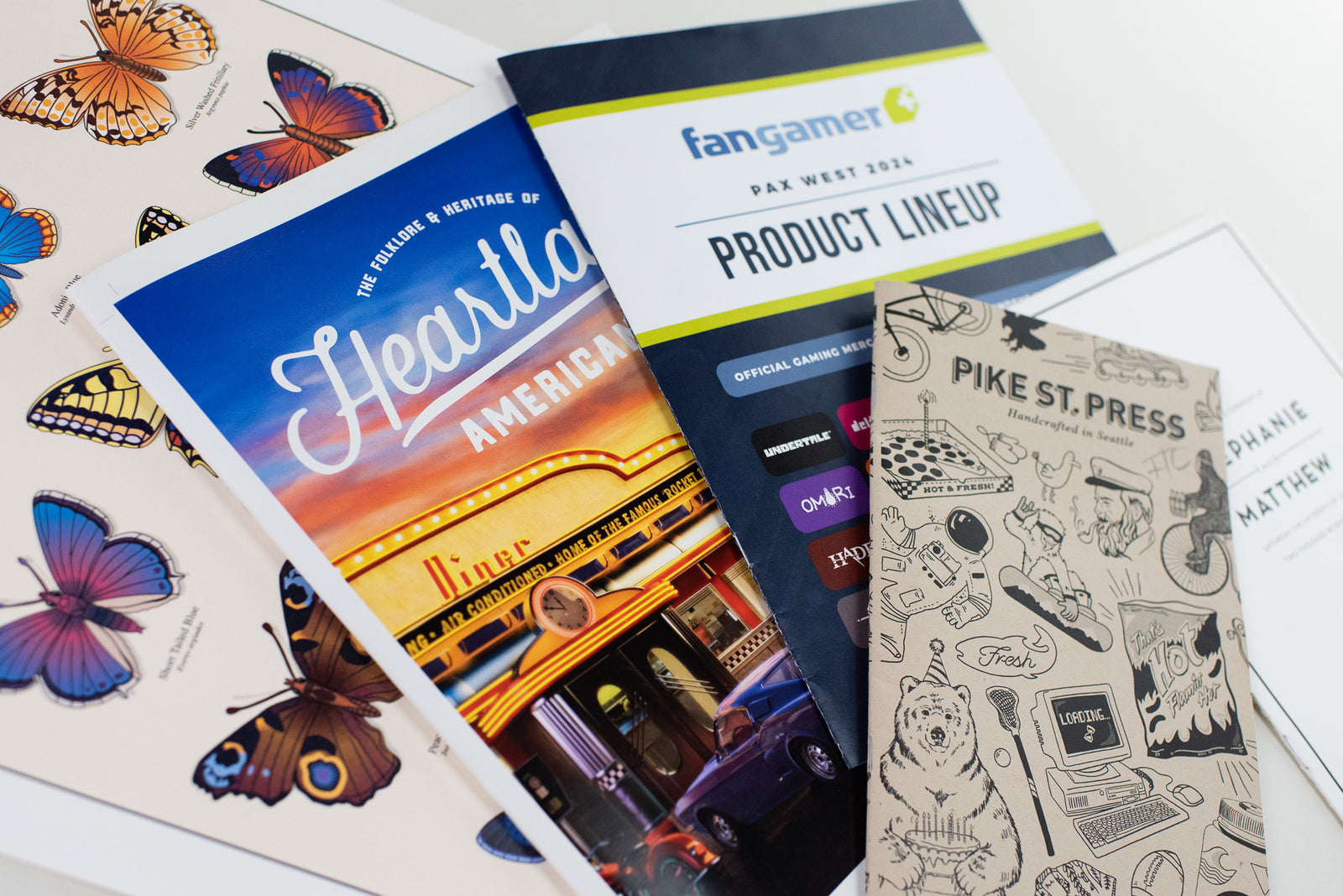 A collection of brochures is laid out on a surface. Among them is one with a butterfly theme, another titled "Heartland American" showcasing a diner image, one from "Pike St. Press" featuring various doodles, and a "Fangamer" brochure displaying their product lineup.