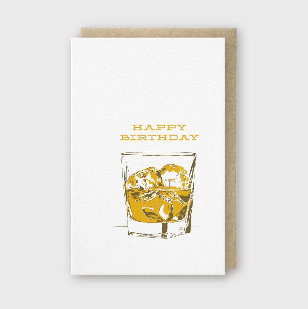 The "Birthday on the Rocks Greeting Card" features a charming "Happy Birthday" message in yellow, created with letterpress techniques. Above, there's a whimsical illustration of whiskey and ice cubes. This handcrafted card from Seattle is accompanied by a brown envelope, set against a light gray background, blending elegance with artisanal charm.