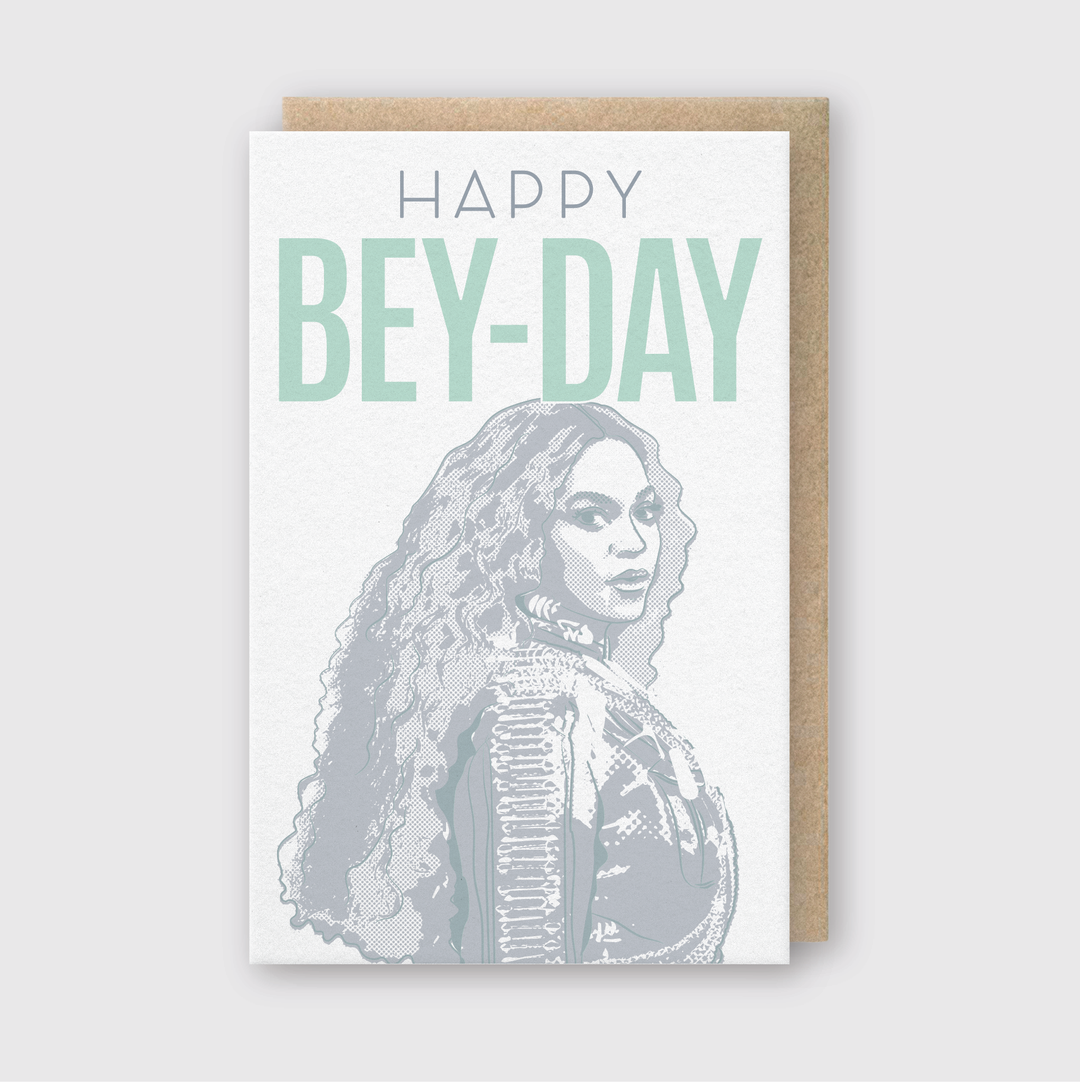 The "Happy Bey-Day" Greeting Card features a striking illustration of a woman with long curly hair in a patterned jacket. Bold text highlights the design. Created using letterpress techniques, this card is paired with a recycled kraft envelope to enhance its eco-friendly appeal against a simple background.