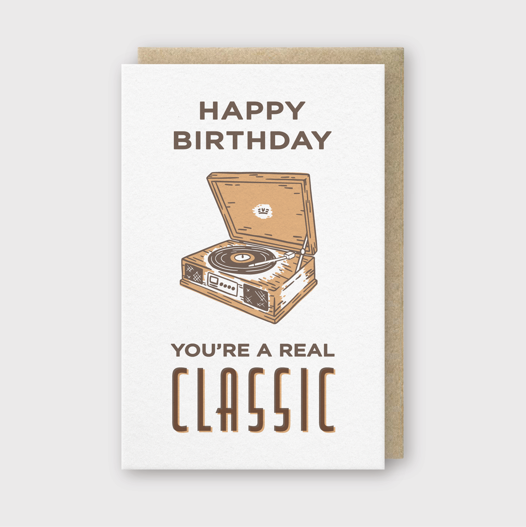 The Happy Birthday Classic Vinyl Greeting Card features a charming illustration of a vintage record player. With "Happy Birthday" at the top and "You're a Real Classic" at the bottom, it perfectly complements your timeless style. The letterpress printing enhances its brown and beige color scheme with added charm.