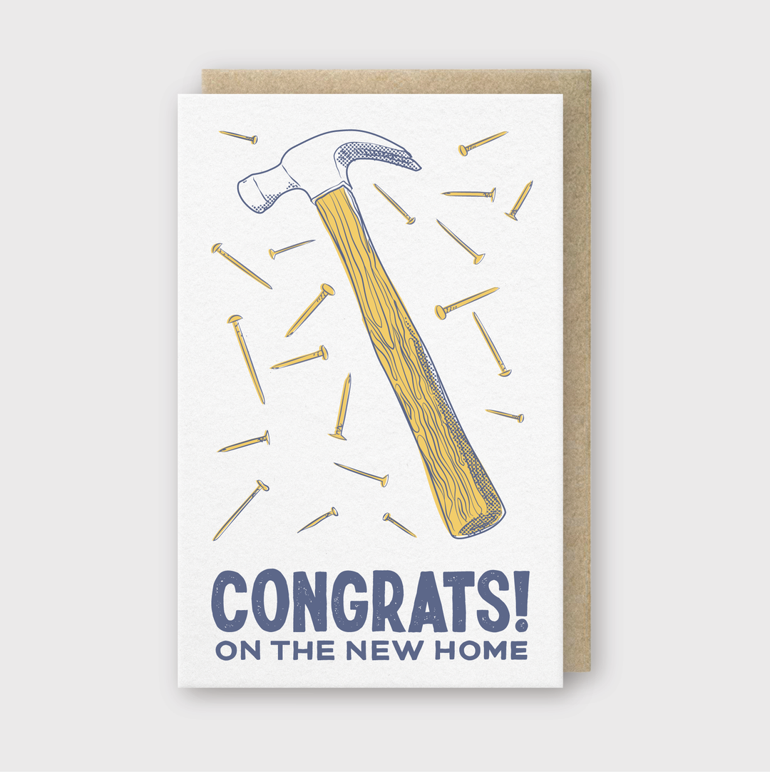 The New Home Hammer Greeting Card is a beautifully crafted letterpress card featuring an illustrated hammer surrounded by scattered nails. It displays the message "CONGRATS! ON THE NEW HOME" and comes with a brown envelope, set against a light gray background—perfect for celebrating new beginnings.
