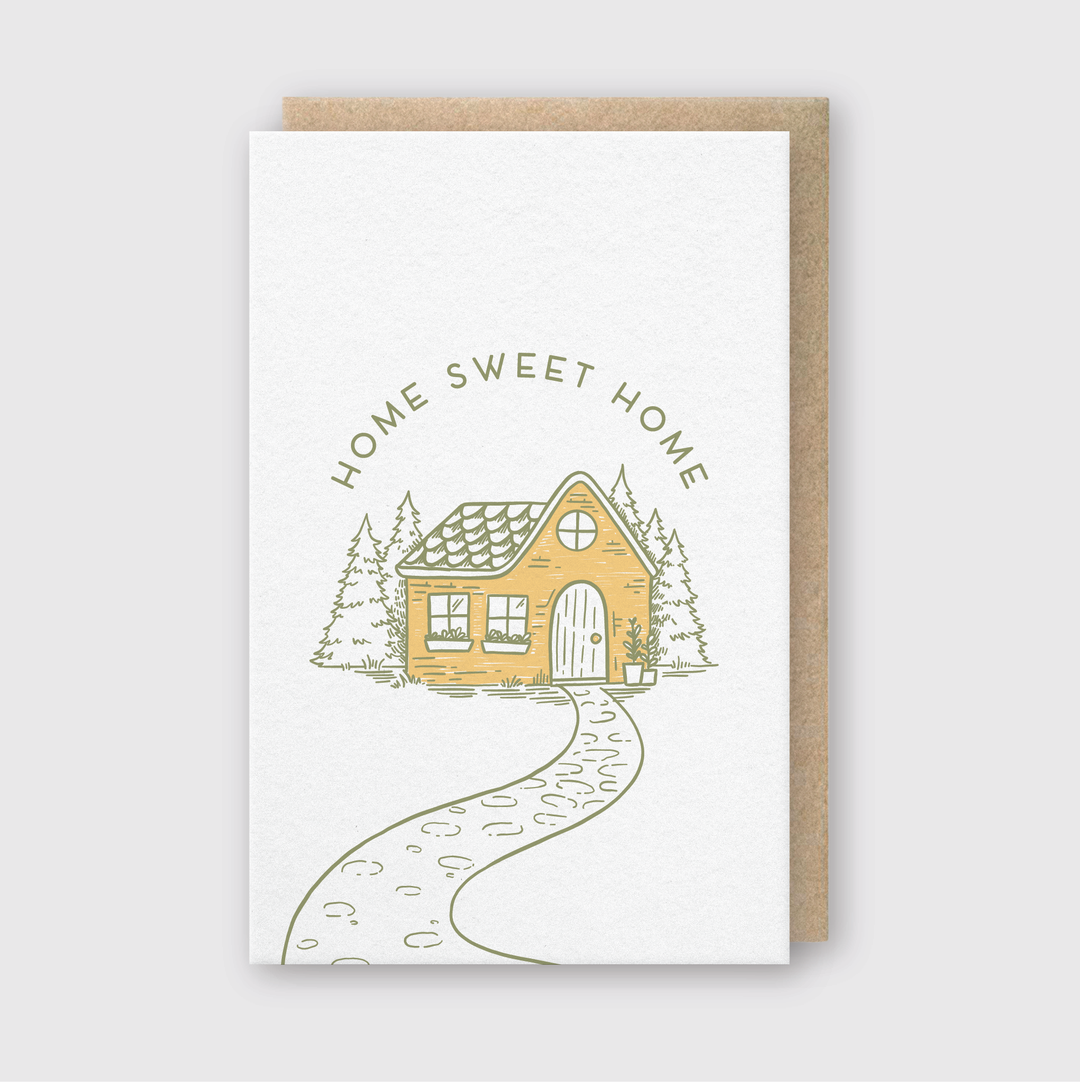Introducing the "Home Sweet Home Gold Greeting Card," an exquisite letterpress card showcasing a charming illustration of a cozy yellow house nestled among trees. A winding pathway leads to the front door, with "Home Sweet Home" beautifully scripted above. It features a brown backing, making it ideal for any occasion.