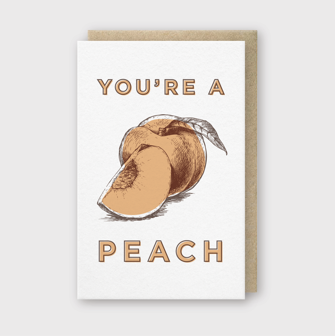The "You're a Peach" Greeting Card showcases an illustration of a whole peach alongside a slice, highlighted by bold brown text that says "You're a Peach." This handcrafted card is made in Seattle and is packaged with a recycled kraft envelope, set against a light gray background.